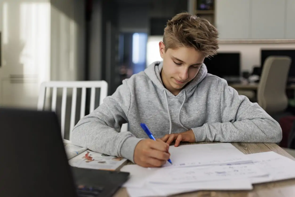 A focused student in a grey hoodie writes notes at a table with a laptop.