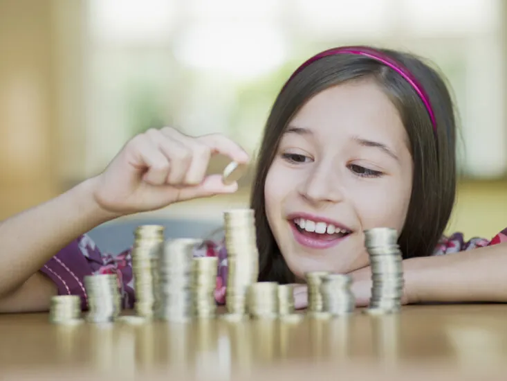 A smiling young girl with a pink headband stacks coins on a table.