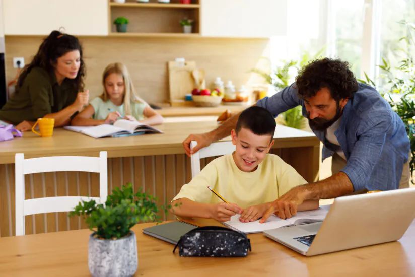 A father helps his smiling son with homework at a kitchen table while the mother assists their