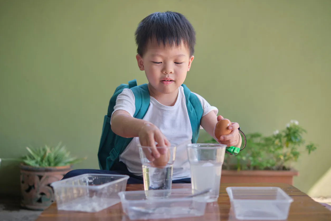 A young boy in a backpack performs an egg science experiment with two cups of water. TITLE