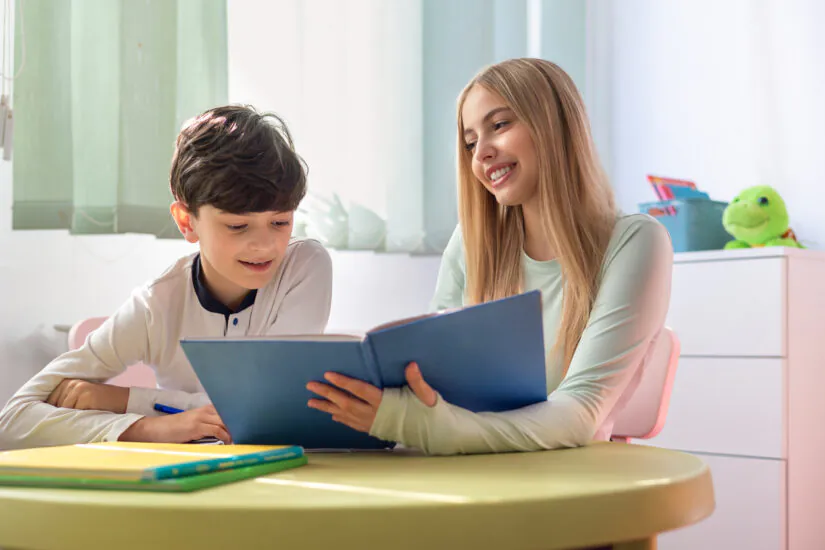 A smiling young woman reads a blue book with a boy at a bright table, engaged