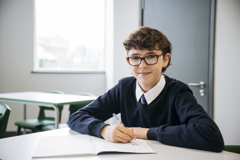 A smiling male student wearing glasses and a uniform writes in a notebook at a classroom desk.
