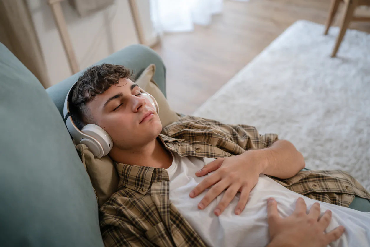 A young man with headphones sleeps peacefully on a teal couch, hands resting on his stomach.