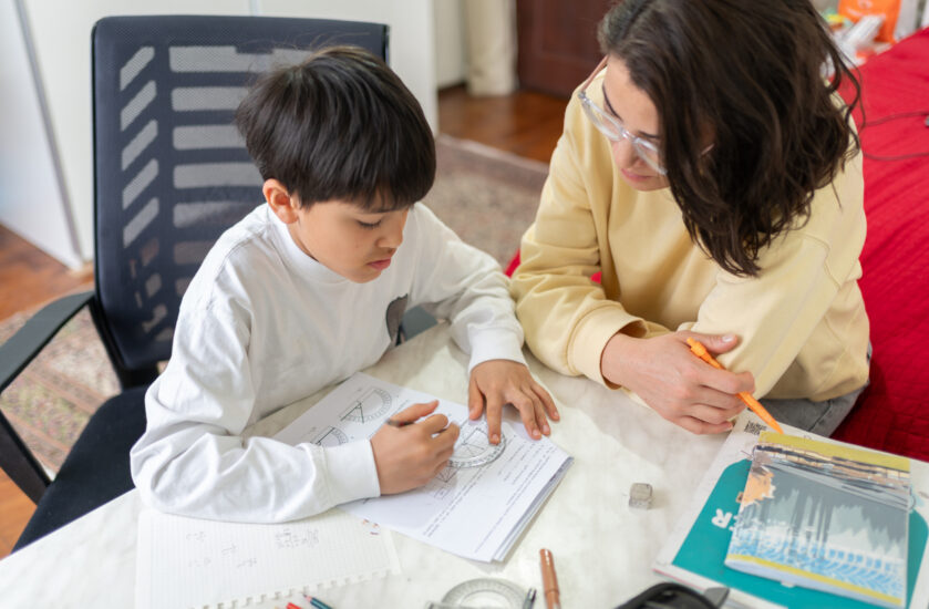 An adult woman tutors a young boy in math, helping him use a protractor.